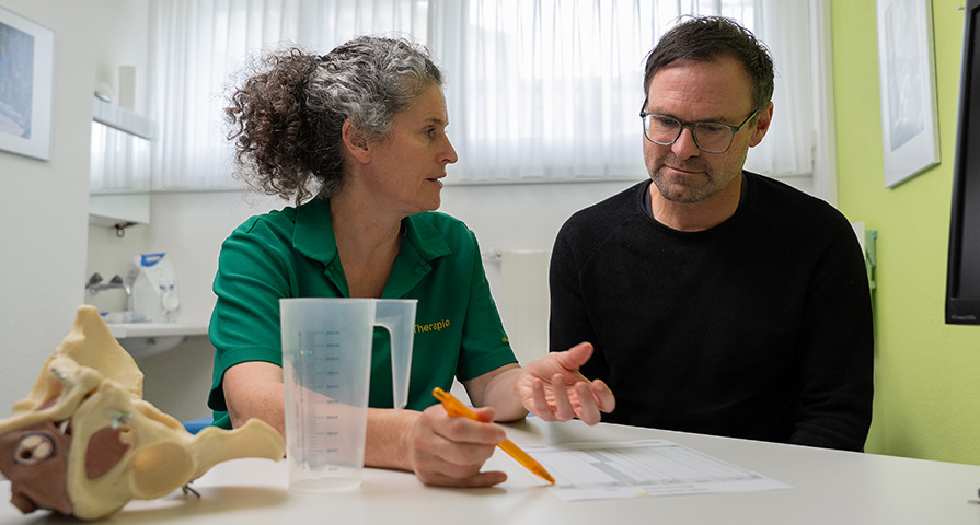 Eine Beckenboden-Physiotherapeutinnen erklärt einem Patienten die Anatomie und Funktion des Beckenbodens